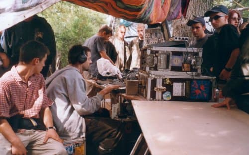 69db of Spiral Tribe (SP23) and Mem Pamal playing live hardware at a free party in the Ardeche, France, in 2000, with onlookers behind the table.
