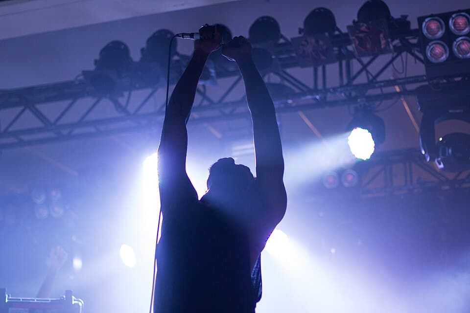 A singer in silhouette holds a microphone above his head under blue stage lights at an Atari Teenage Riot concert at Amphi festival in Cologne, 2013.