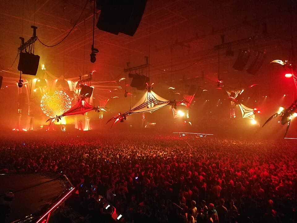 A packed indoor arena lit in red at Qlimax 2017, with lit canopies and a glowing sphere hanging over the crowd.